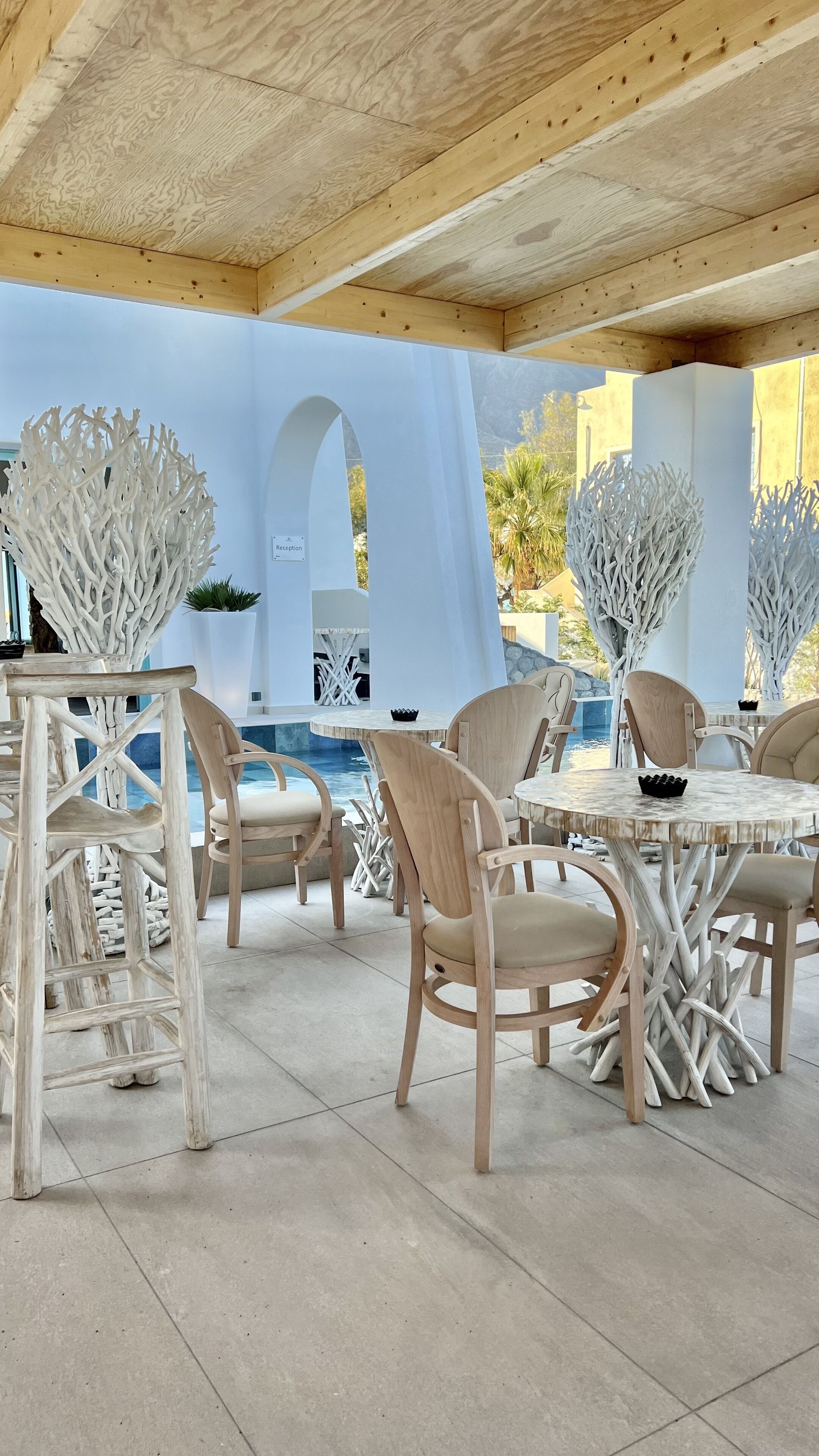 An outdoor seating area with wooden chairs and tables, a pool, and white architectural features.