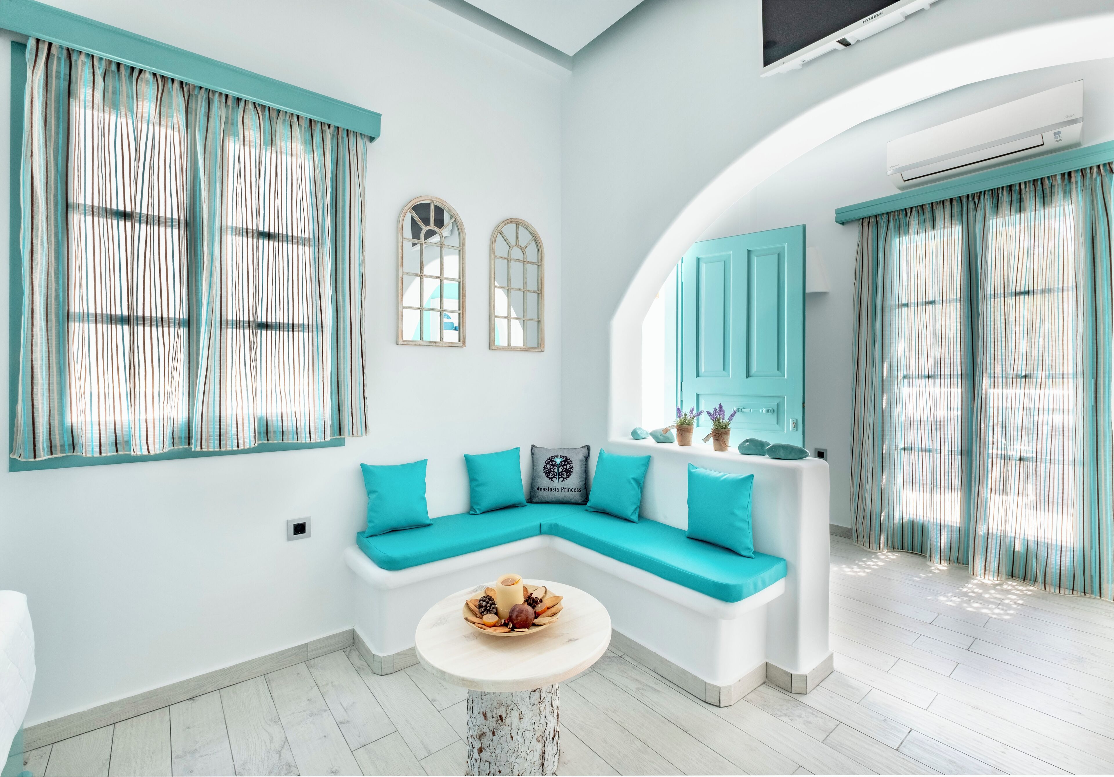 A modern living room with a white sectional sofa, teal cushions, a round coffee table with fruit, and a large window with striped curtains.