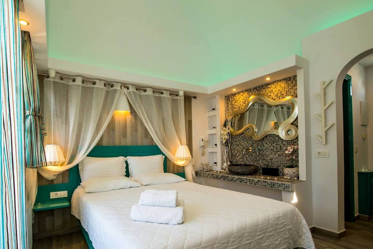 Een netjes opgemaakt bed met witte lakens en twee handdoeken, een nachtkastje met een lamp, een grote spiegel met decoratieve lijst, en uitzicht op een betegelde muur en een groene deur.