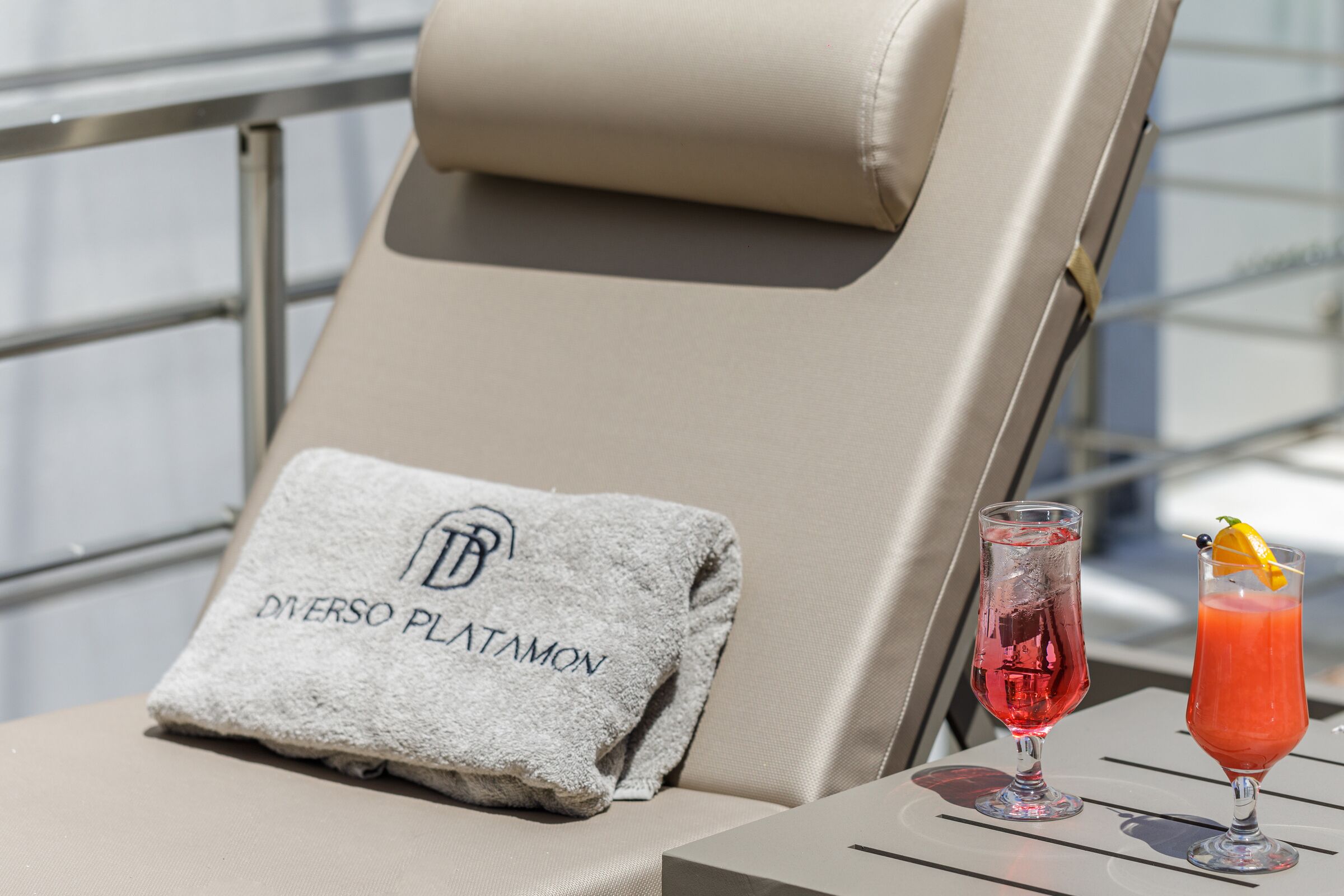 A lounge chair with a towel labeled "DIVERSO PLATAMON" and a glass of red cocktail with a garnish on a table.