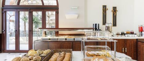 A breakfast buffet with various breads and pastries, a knife and spoon on a cutting board, and a view of palm trees through large windows.