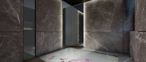 Un baño moderno con paredes de mármol oscuro, cabina de ducha de vidrio y un suelo cubierto de pétalos de rosa.