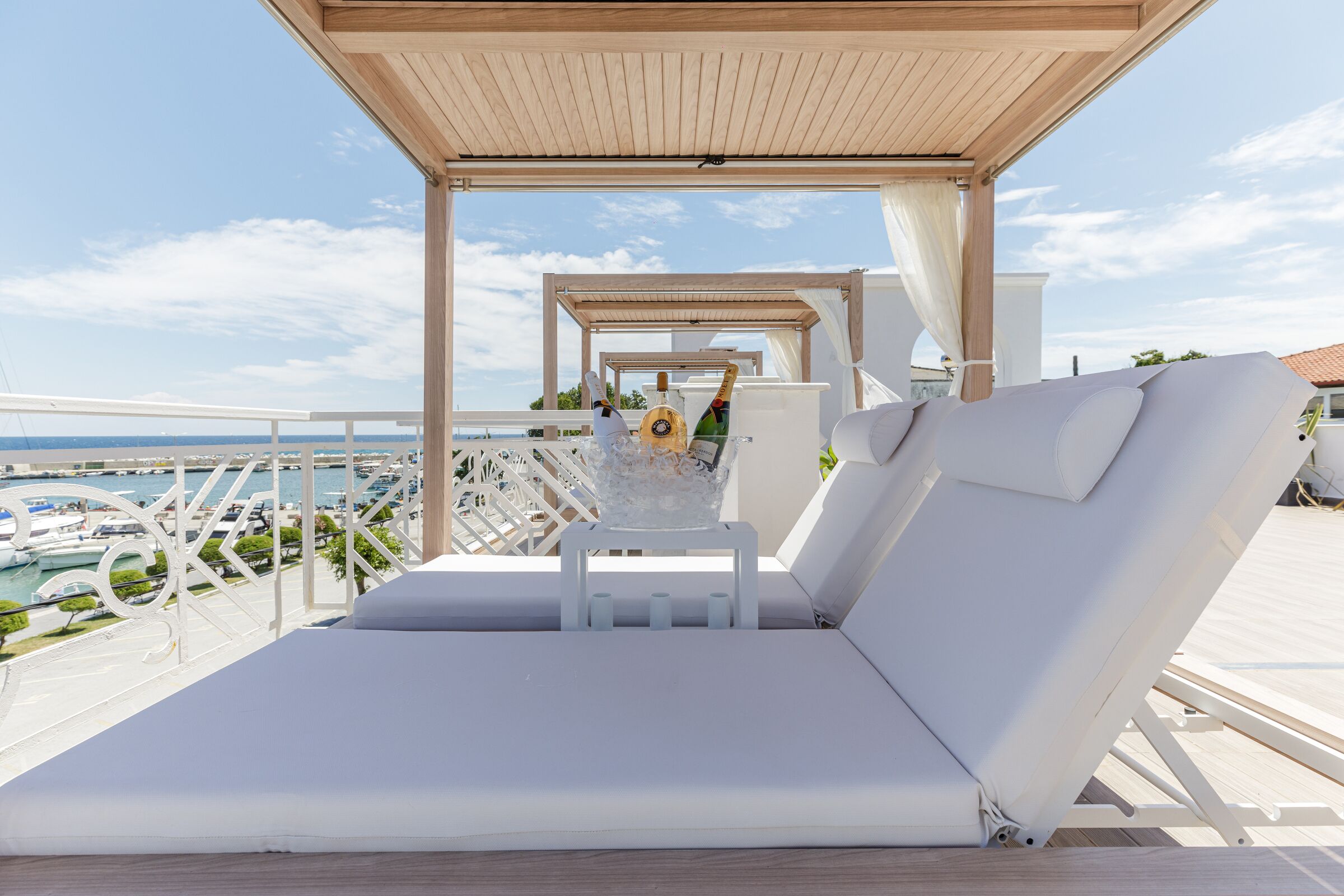 A sunlit outdoor lounge area with white cushioned seating, a wooden pergola, and a view of a marina.