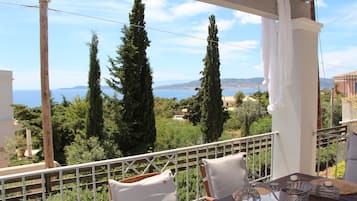 A balcony with a view of the sea, tall cypress trees, and a table with cushions and candles.