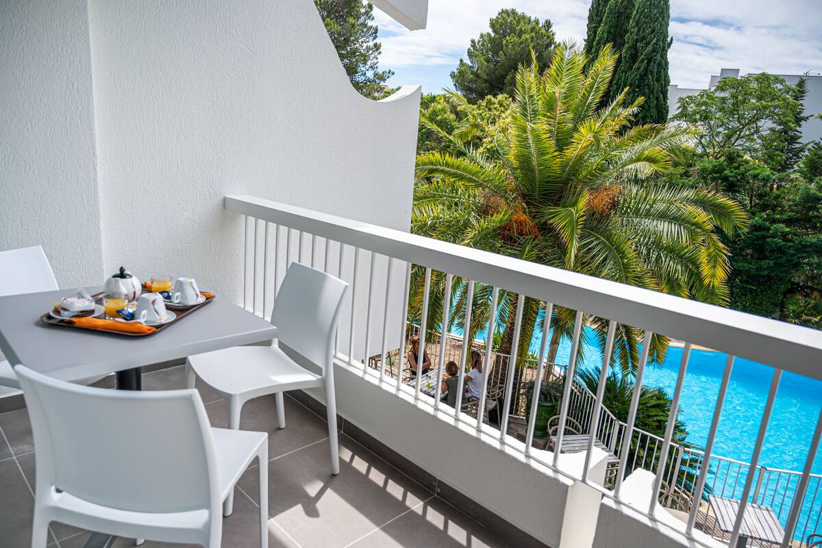 A balcony with white chairs and a table set for breakfast, overlooking a swimming pool and lush greenery.