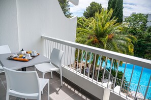 A balcony with white chairs and a table set for breakfast, overlooking a swimming pool and lush greenery. - Hôtel Le Quetzal (La Grande-Motte)