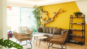 Une pièce lumineuse avec un mur jaune, un canapé, des fauteuils en osier, une petite table et une bibliothèque.