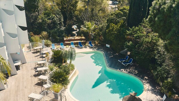 Vue aérienne d’un complexe hôtelier doté d’une piscine entourée de chaises longues et de parasols, d’une terrasse en bois et d’une végétation luxuriante.