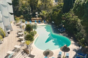 An aerial view of a resort with a swimming pool surrounded by lounge chairs and umbrellas, a wooden deck, and lush greenery. - Hôtel Le Quetzal (La Grande-Motte)