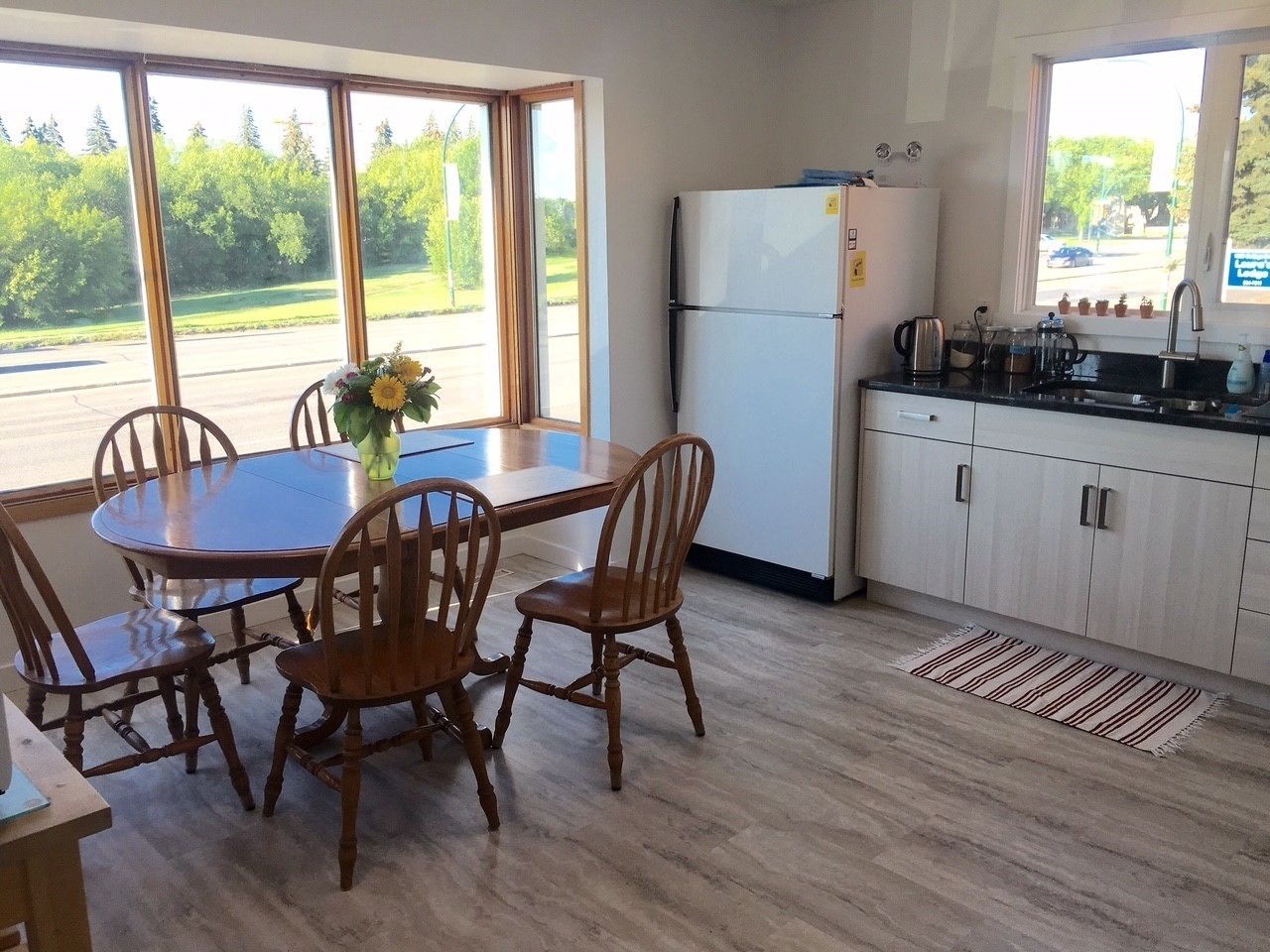 A kitchen with a dining table and chairs, a refrigerator, and a window with a view of trees.