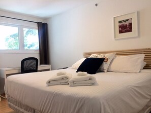 A neatly made bed with white linens and two folded towels, a wooden headboard, a desk with a chair, and a window with curtains.