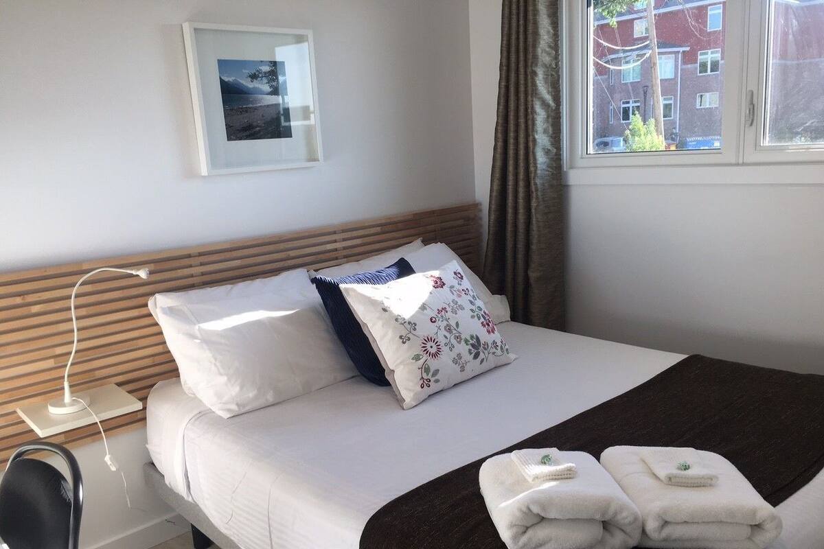 A neatly made bed with white linens, a wooden headboard, and a floral pillow. There are two folded towels on the bed and a bedside table with a lamp.