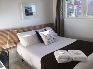 A neatly made bed with white linens, a wooden headboard, and a floral pillow. There are two folded towels on the bed and a bedside table with a lamp.