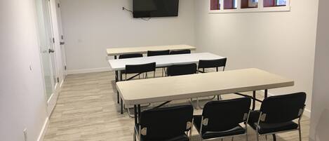 A conference room with tables and chairs, a flat-screen TV, and windows with wooden frames.