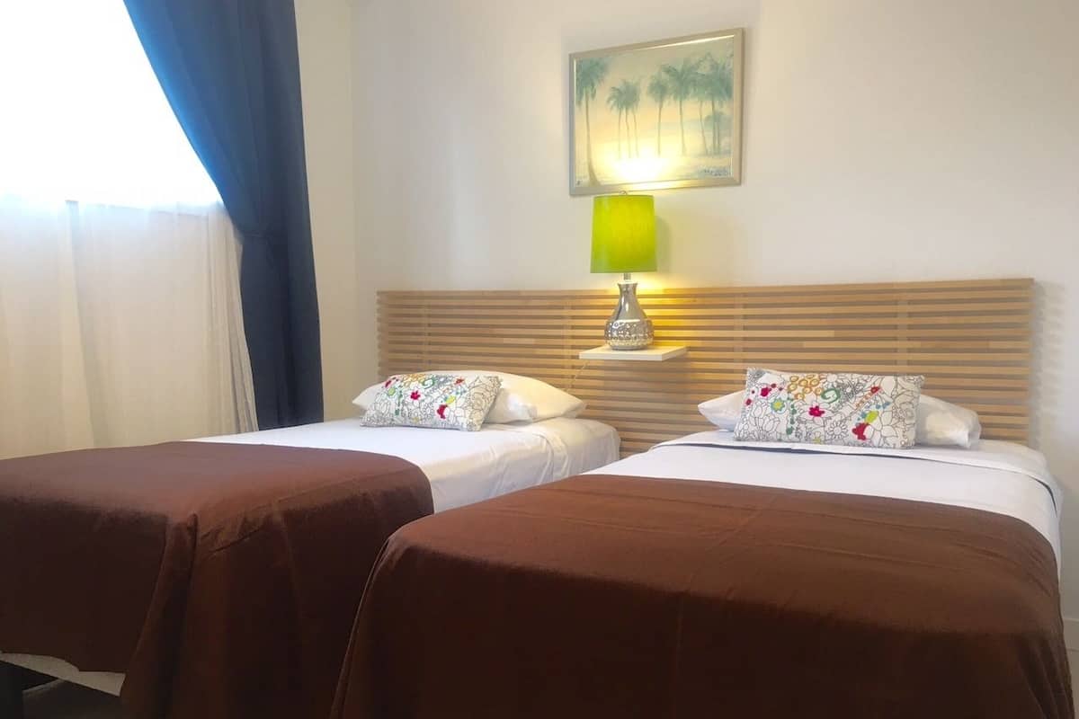 A hotel room with two beds, each with a brown bedspread and floral pillows. There is a nightstand with a lamp between the beds. A painting of palm trees hangs on the wall above the nightstand.