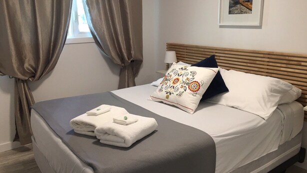 Una cama bien hecha con una colcha gris, almohadas blancas y una almohada decorativa. Hay toallas blancas dobladas sobre la cama.
