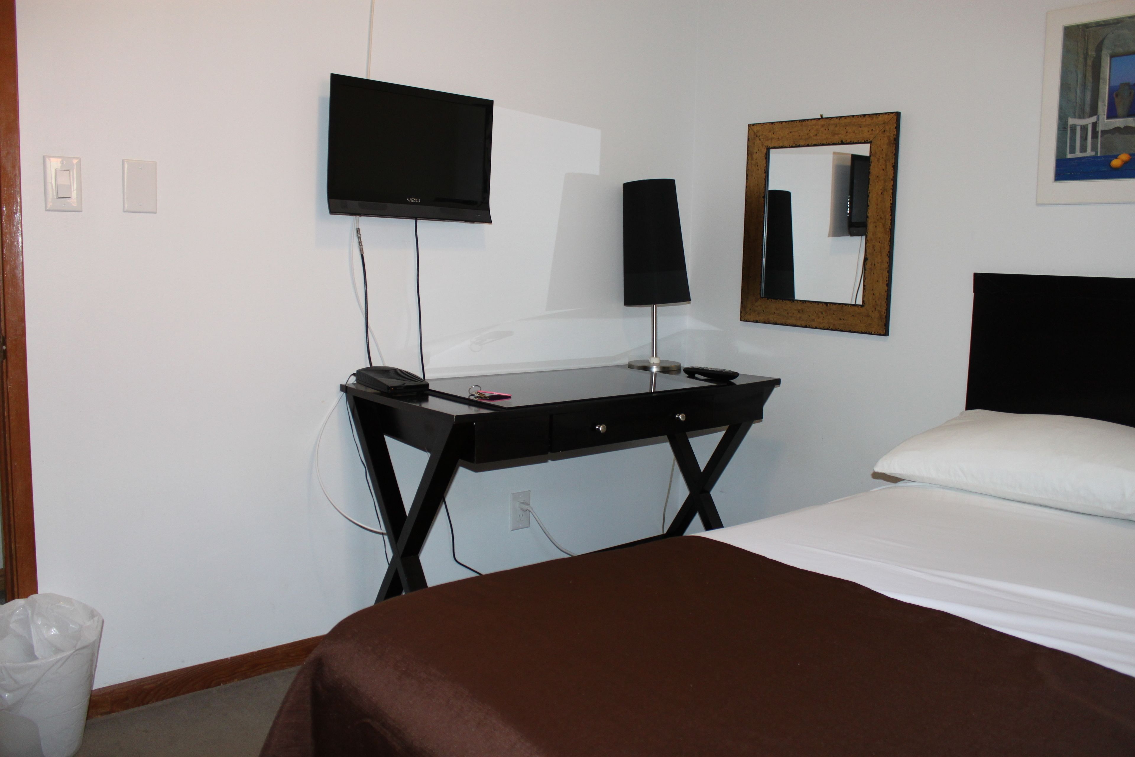 A hotel room with a bed, a desk with a computer, a TV, a mirror, and a trash bin.
