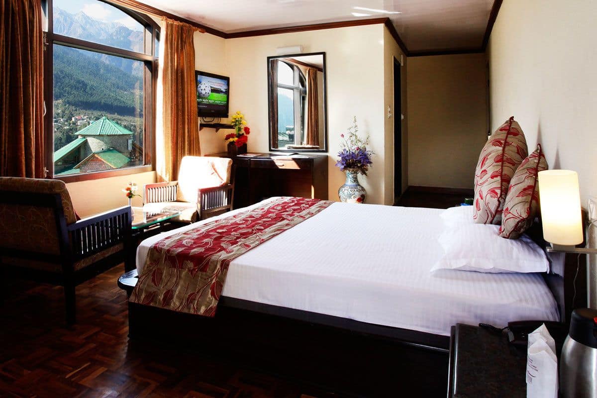 A hotel room with a large bed, a desk with a chair, a TV, a window with curtains, and a vase with flowers.