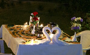A table set outdoors with a white tablecloth, candles, a vase with flowers, and a heart-shaped towel arrangement.