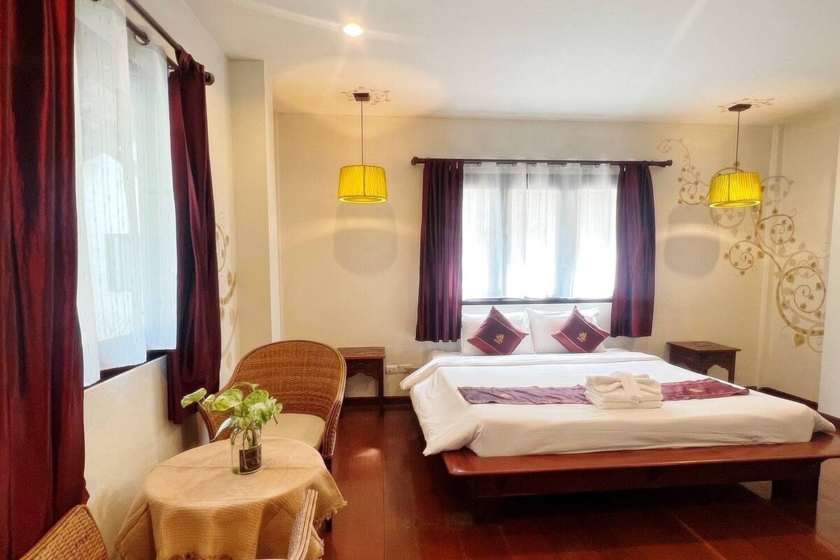 Une chambre d’hôtel avec un grand lit, deux tables de chevet, une petite table ronde avec une plante, des chaises en osier et deux lampes suspendues.
