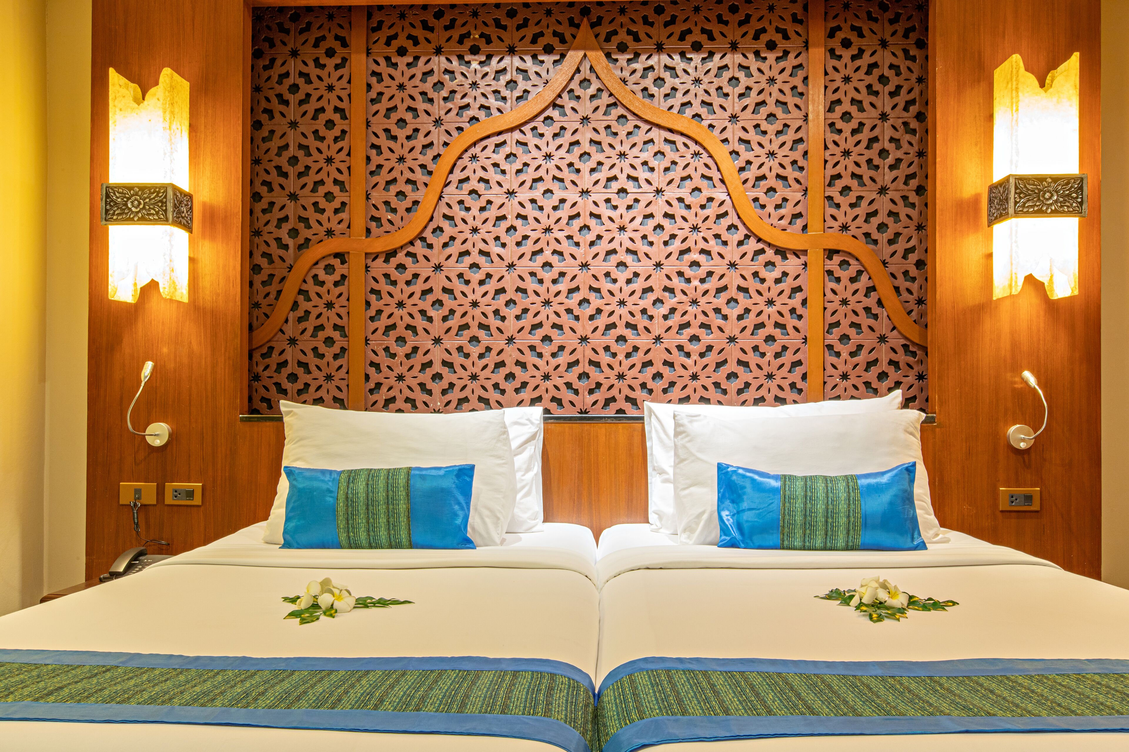 A double bed with white and blue bedding, two wall-mounted lamps, and a decorative wooden headboard with intricate patterns.