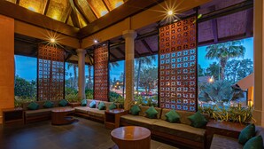 A spacious lounge area with multiple seating options, including sofas and a round table, and large windows offering views of palm trees and a sunset. - Rawai Palm Beach Resort (Rawai)