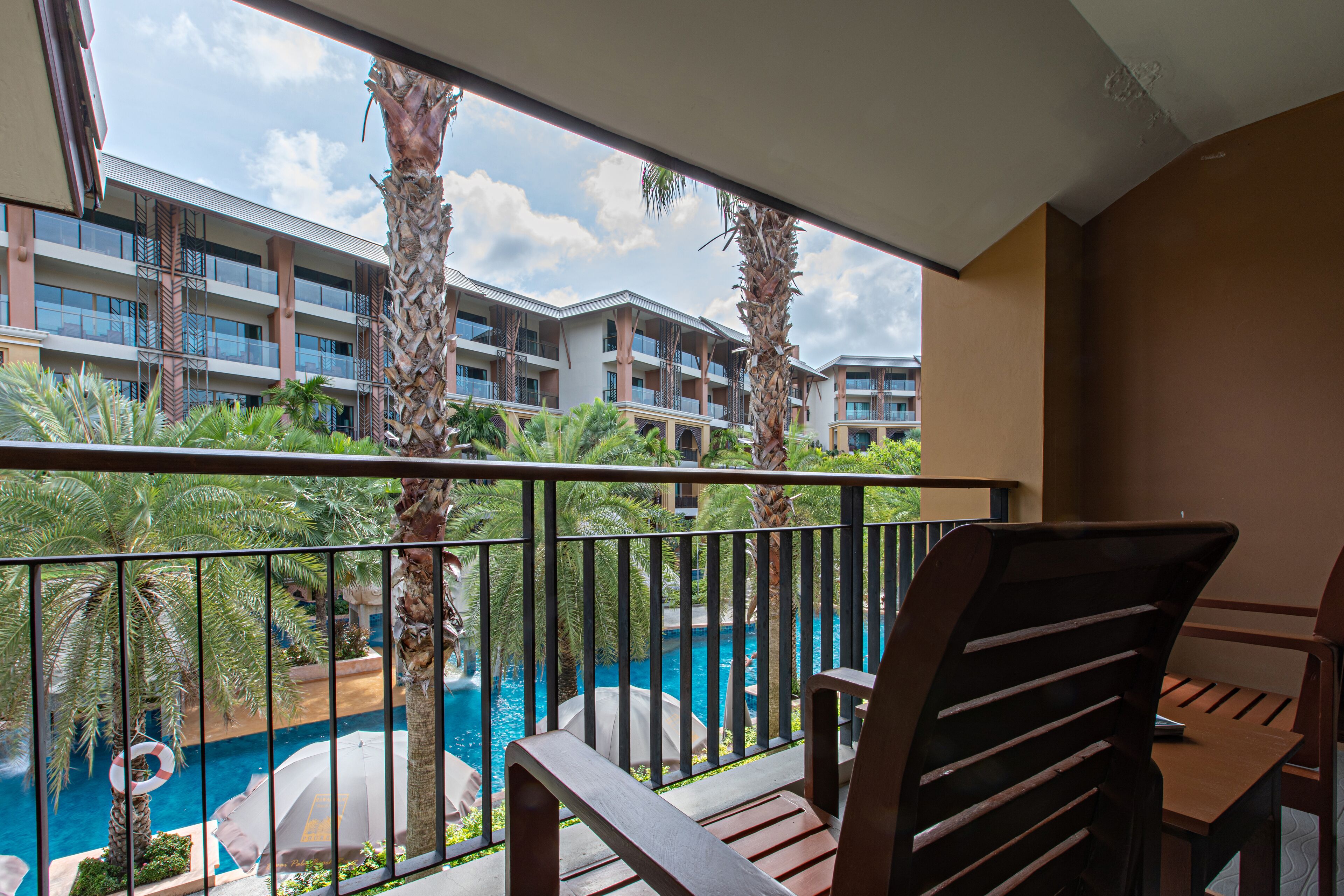 A balcony with a view of a swimming pool, palm trees, and a hotel building.
