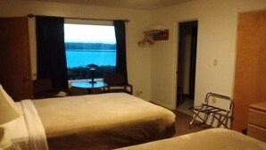 A hotel room with two beds, a small table, and a chair. There is a window with a view of a body of water and trees. - Bay Bridge Motel (North Bend)