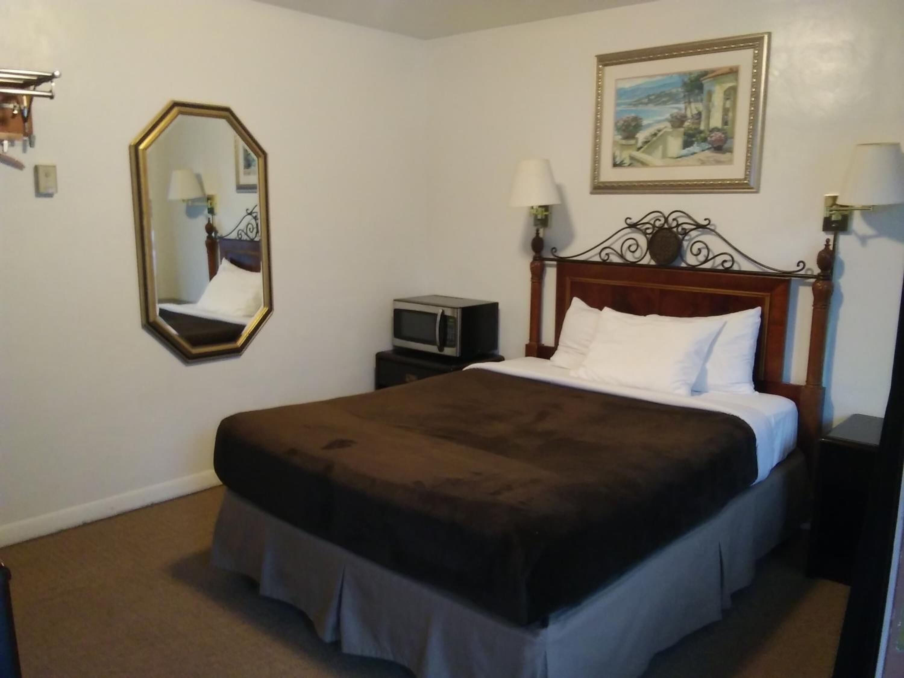 A hotel room with a large bed, a wooden headboard, a nightstand, a small table, a mirror, and a painting on the wall.
