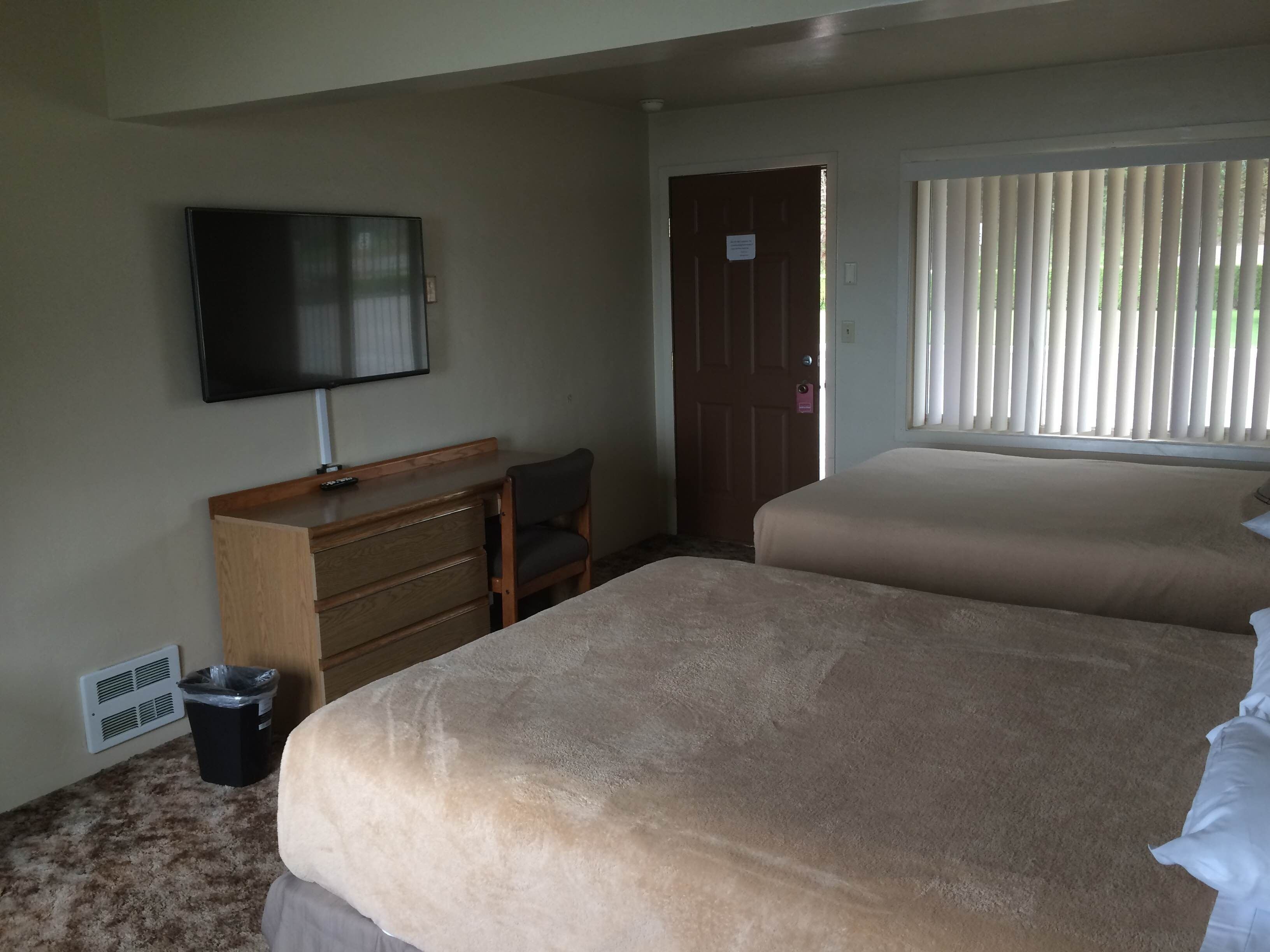 Ein Hotelzimmer mit zwei Betten, einem Schreibtisch, einem Sessel, einem Fernseher, einem Fenster mit Jalousien und einer Tür.