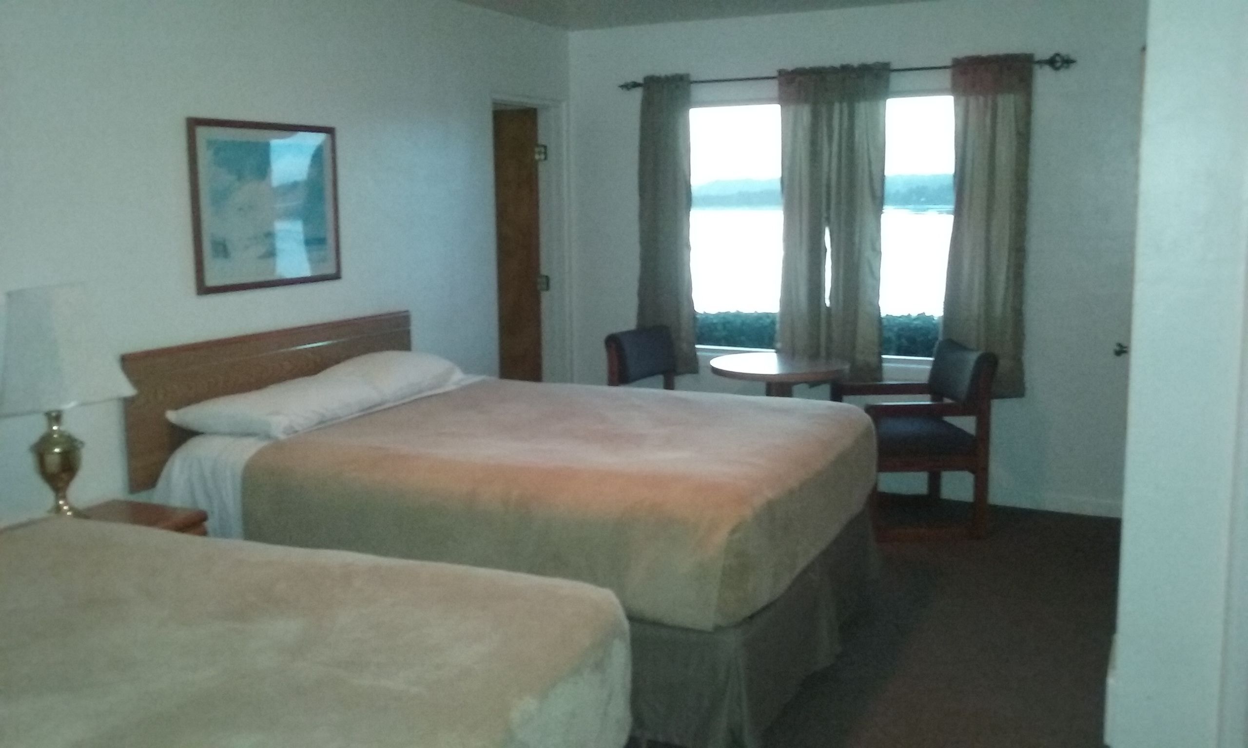 A hotel room with two beds, a small table with a chair, a lamp, and a window with curtains.