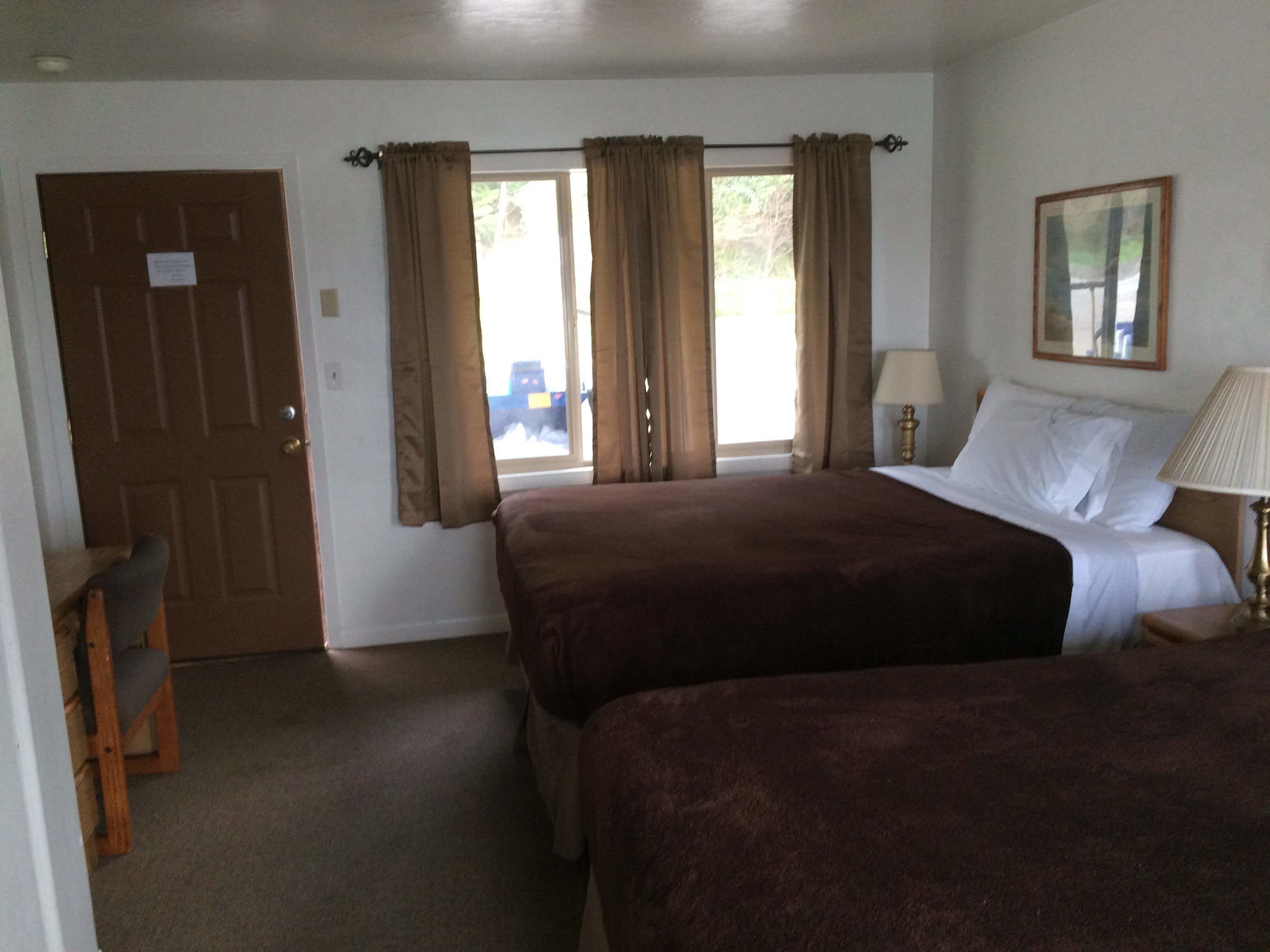 A hotel room with two beds, a wooden chair, a desk, a lamp, and a window with curtains.