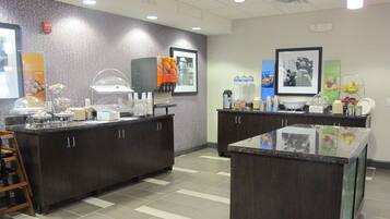 A self-service breakfast area with a variety of food and drink options, including a coffee station and a fruit display.