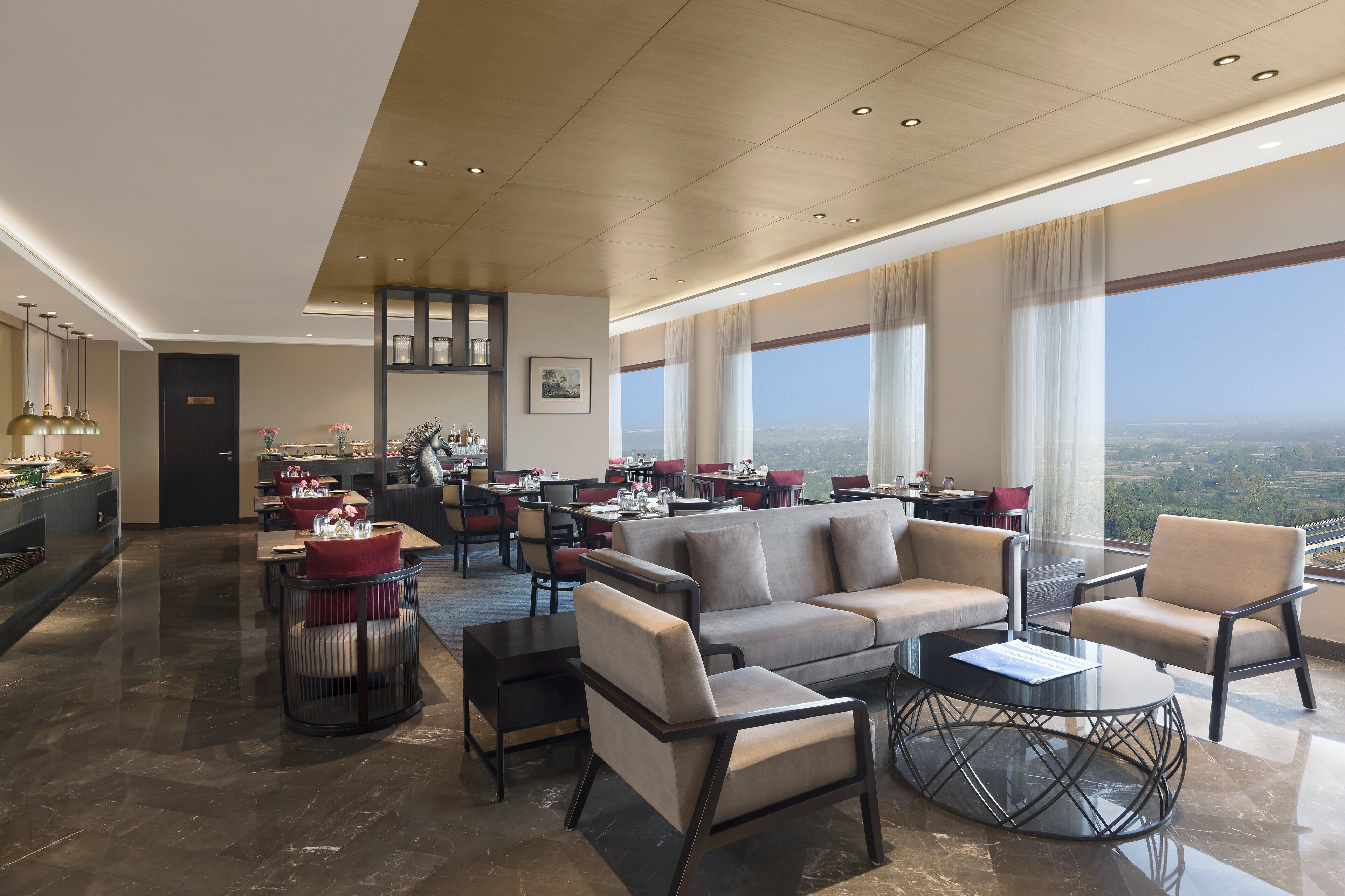 A spacious lounge area with a sofa, armchairs, and a coffee table. There is a bar area with a counter and stools. Large windows provide a view of the cityscape.