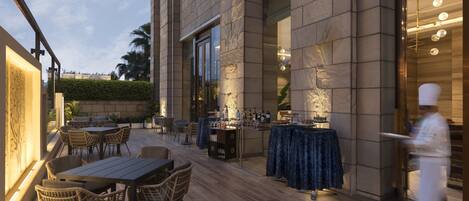 An outdoor dining area with wicker chairs and tables, a bar area with a blue tablecloth, and a chef in uniform.
