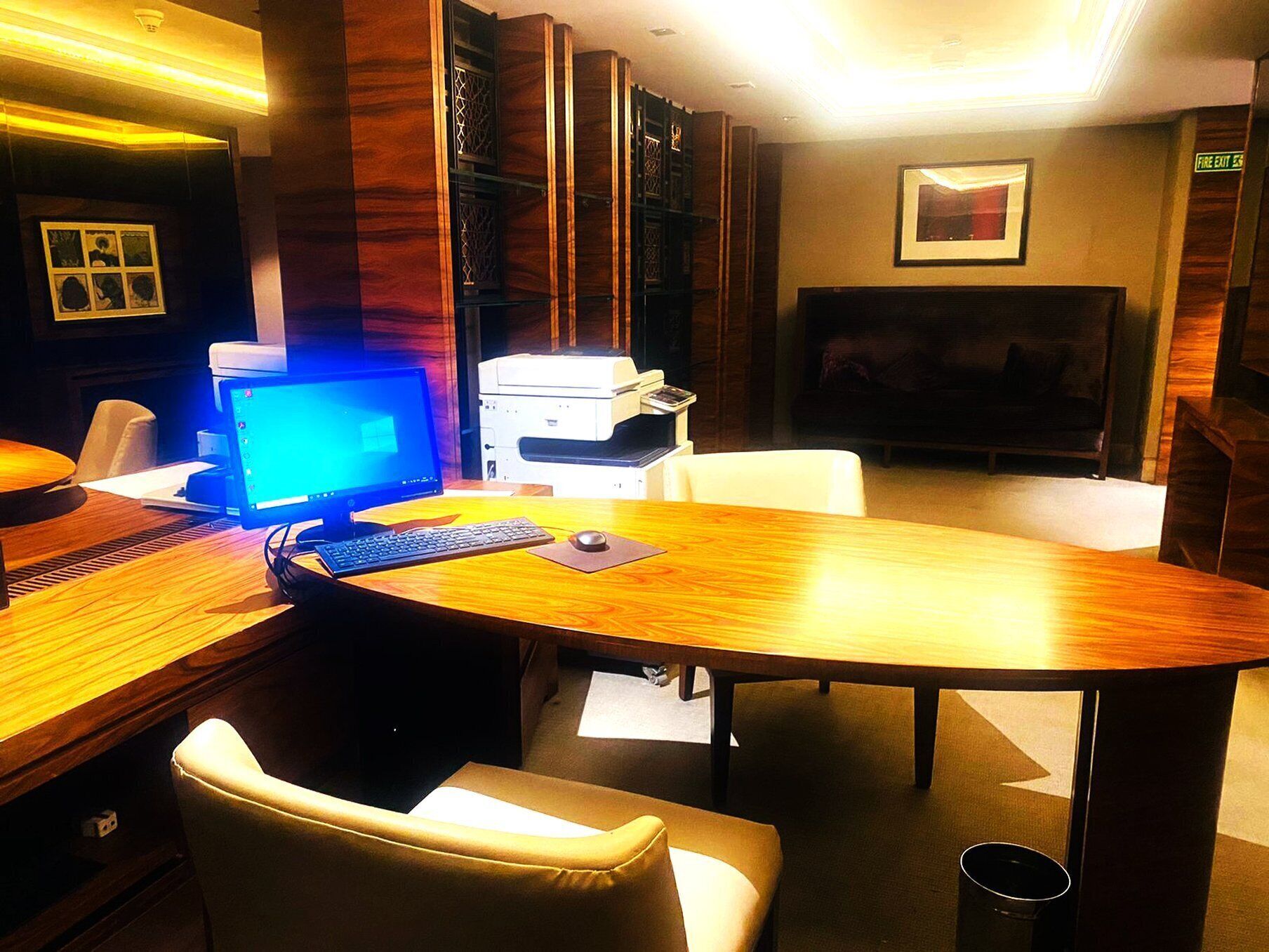 A hotel room with a wooden desk, a computer, a printer, a TV, and a sofa.