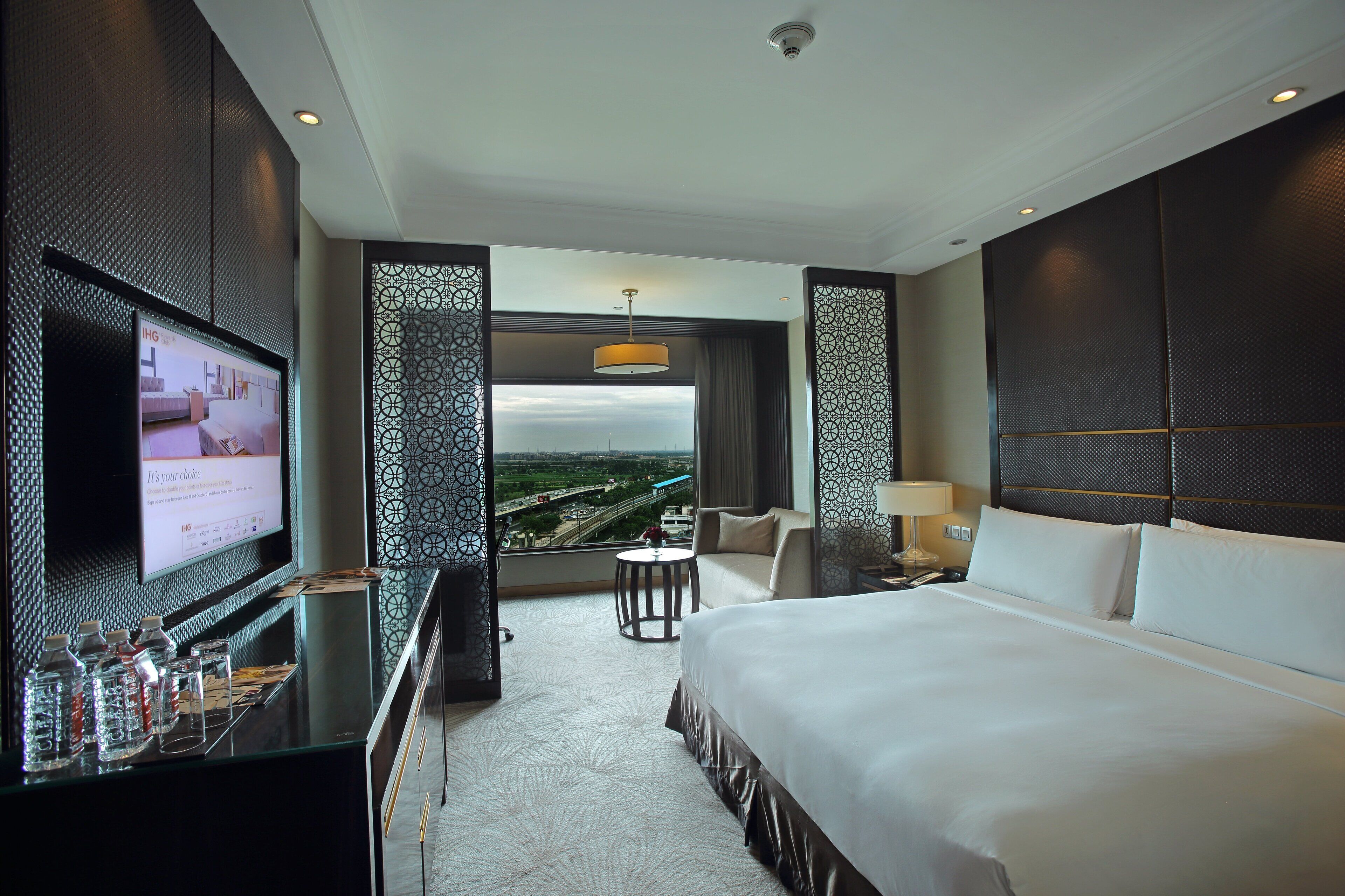 A hotel room with a large bed, a TV, a small table with a lamp, and a view of the cityscape.
