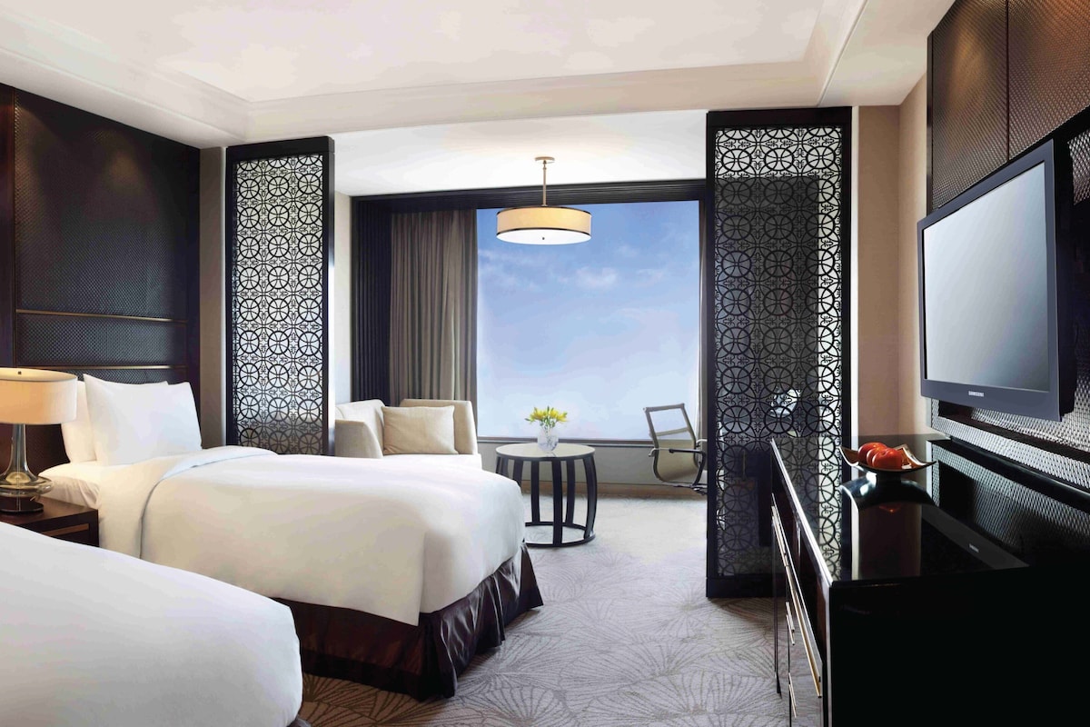 A hotel room with two beds, a desk, a TV, and a balcony with a view.