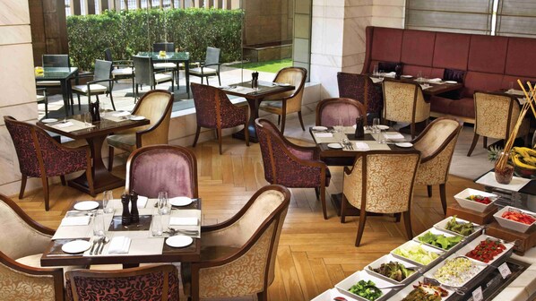 A hotel dining area with multiple tables and chairs, a buffet with various food options, and a view of the outdoor garden.