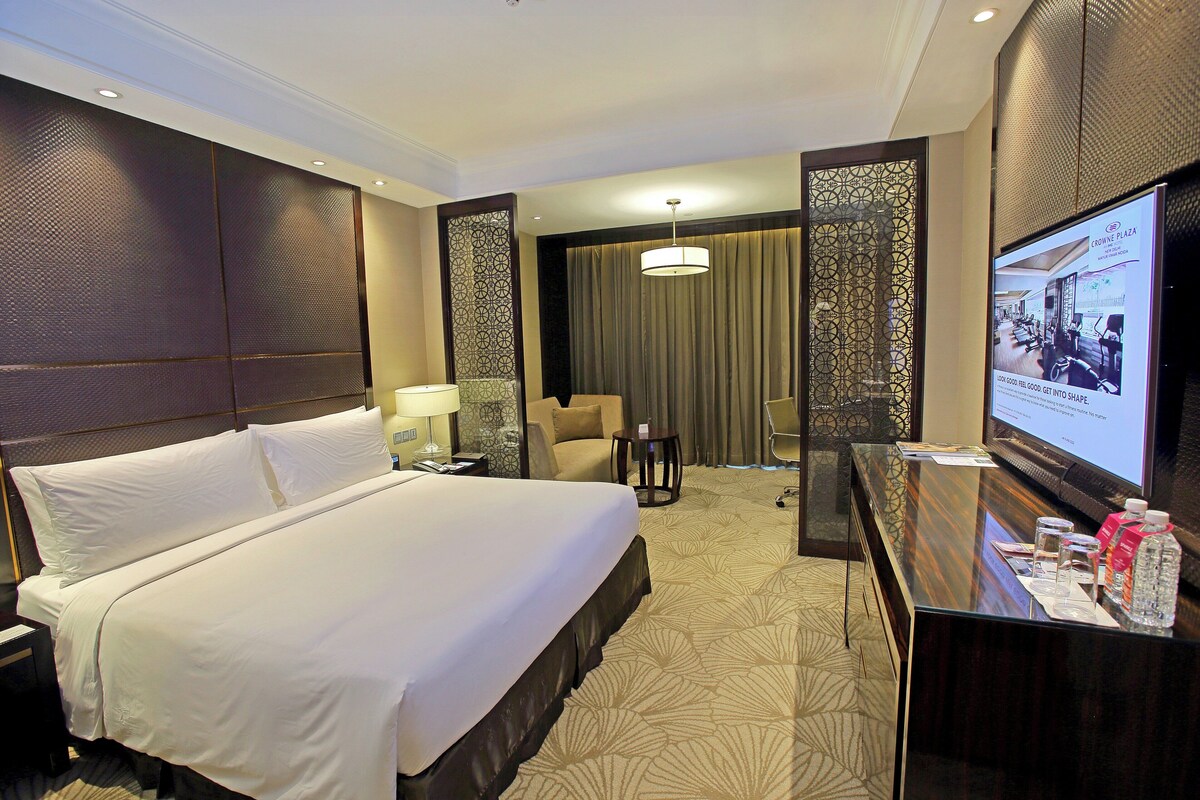A hotel room with a large bed, a desk with a computer, a chair, a TV screen, and a decorative wall panel.