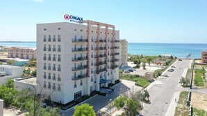 A multi-story hotel with a prominent red construction crane, a clear view of the sea, and a parking area in front.