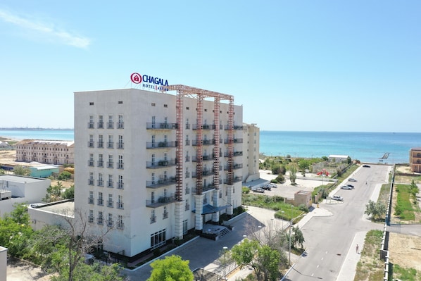 A multi-story hotel with a prominent red construction crane, a clear view of the sea, and a parking area in front.