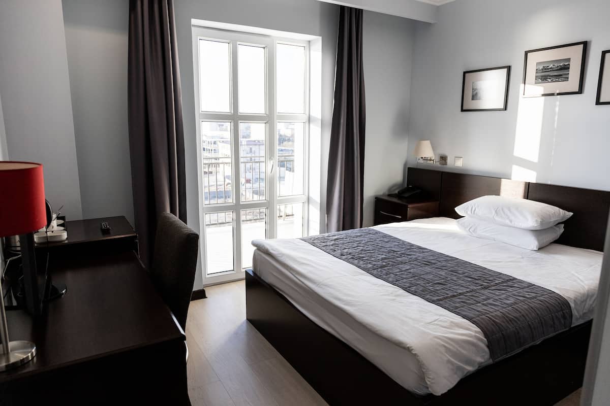 A hotel room with a bed, a desk, a chair, a nightstand, a lamp, and a window with curtains.