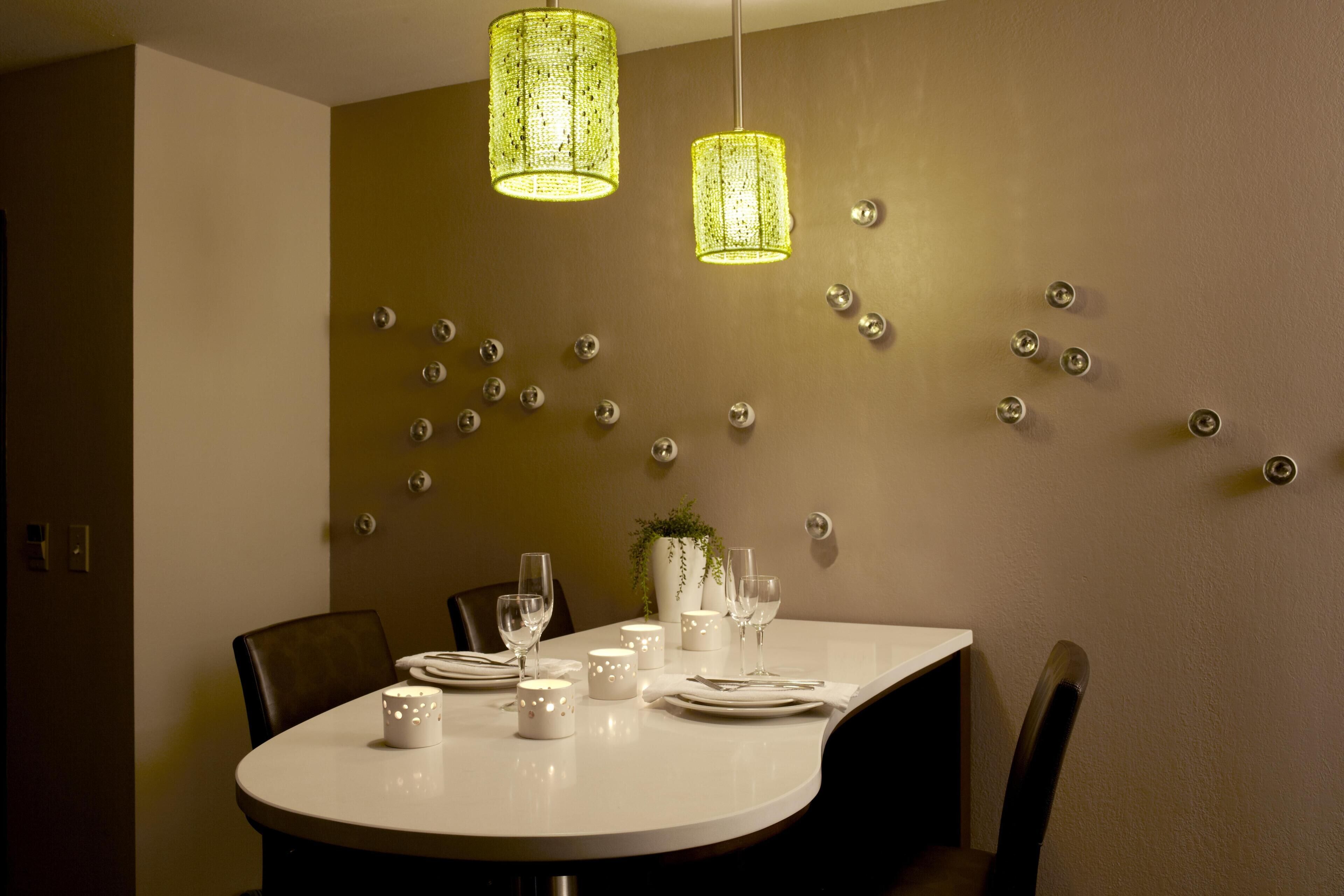Un coin repas avec une table ronde dressée pour deux personnes, deux lampes suspendues et un mur orné de motifs métalliques décoratifs.