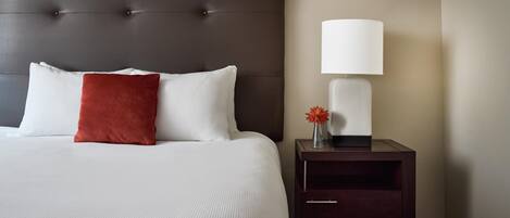 A neatly made bed with a dark headboard, white and red pillows, a bedside table with a lamp, and a vase with flowers.