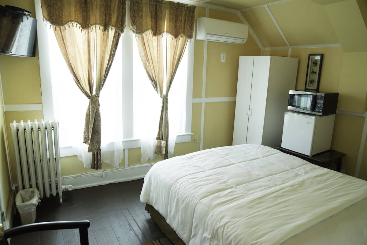 Une chambre à coucher avec un lit, un radiateur, une armoire blanche, un four à micro-ondes et une fenêtre avec des rideaux.