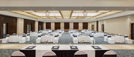 Un amplio salón de conferencias con varias mesas y sillas dispuestas en filas, suelo alfombrado y un techo alto con tragaluces.