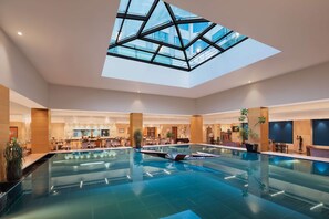 A spacious indoor swimming pool with a skylight, surrounded by seating areas and a bar. - Radisson Blu Hotel Ranchi (Ranchi)
