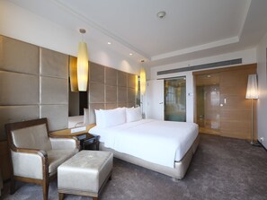A modern hotel room with a large bed, a sitting area with a chair and ottoman, a bedside table, and a walk-in closet.