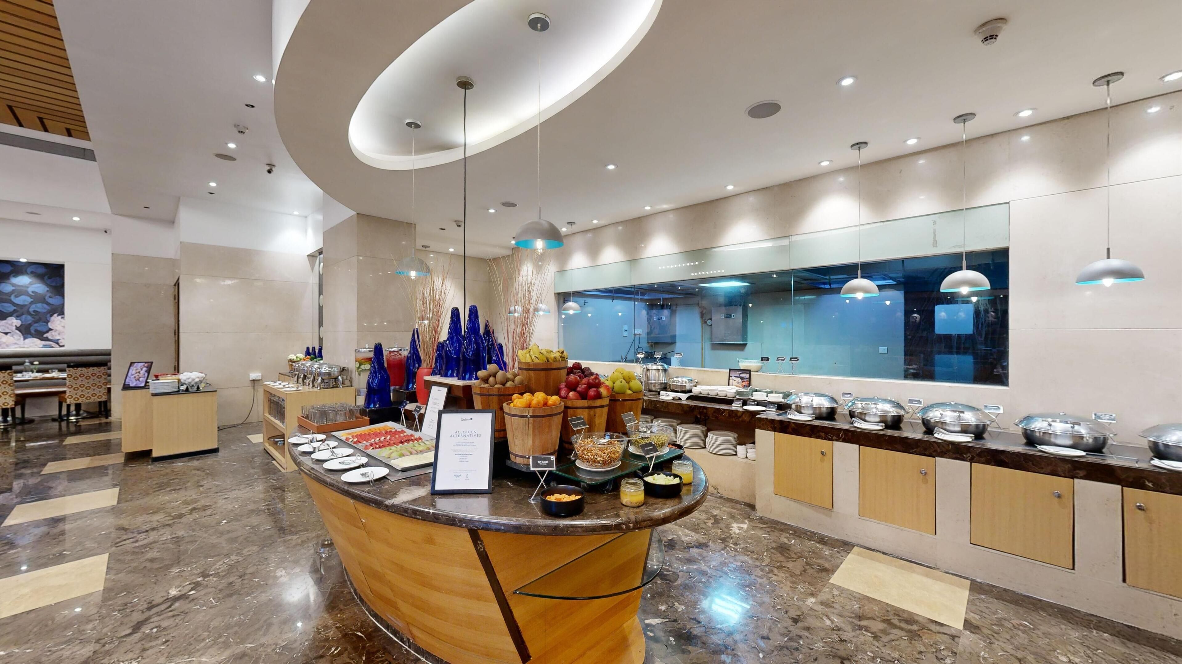 En moderne hotellobby med en receptionsdisk i marmor, et buffetområde med forskellige retter, og et stort spejl der reflekterer køkkenområdet.